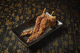 On a dark plate, three crispy fried chicken wings are artfully arranged. The background is covered in an intricate pattern of elegant gold calligraphy on a textured black surface, creating a sophisticated atmosphere.