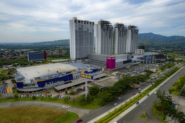 Aerial view of a large commercial complex featuring a prominent IKEA store and adjacent high-rise buildings. The complex is surrounded by roads, parking lots filled with cars, and green landscapes. The AEON shopping mall is also visible with its distinct pink signage. In the background, rolling hills and residential areas extend towards the horizon under a partly cloudy sky.