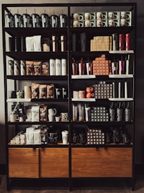 A neatly organized store shelf displaying various branded merchandise including coffee mugs, tumblers, bags of coffee beans, and cups. The products are arranged in a visually appealing manner with different collections and themes.