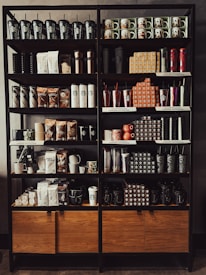 A neatly organized store shelf displaying various branded merchandise including coffee mugs, tumblers, bags of coffee beans, and cups. The products are arranged in a visually appealing manner with different collections and themes.