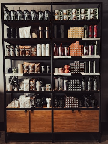 A neatly organized store shelf displaying various branded merchandise including coffee mugs, tumblers, bags of coffee beans, and cups. The products are arranged in a visually appealing manner with different collections and themes.