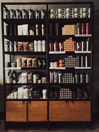 A neatly organized store shelf displaying various branded merchandise including coffee mugs, tumblers, bags of coffee beans, and cups. The products are arranged in a visually appealing manner with different collections and themes.