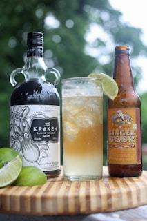 A bottle of dark rum with an intricate nautical design label stands next to a bottle of ginger beer on a wooden surface. Between them is a tall glass filled with a mixed drink, ice cubes, and garnished with a wedge of lime. Additional lime wedges rest on the wooden surface. The background is a blurred green, suggesting an outdoor setting.