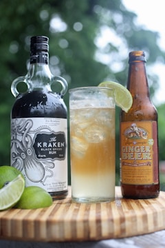 A bottle of dark rum with an intricate nautical design label stands next to a bottle of ginger beer on a wooden surface. Between them is a tall glass filled with a mixed drink, ice cubes, and garnished with a wedge of lime. Additional lime wedges rest on the wooden surface. The background is a blurred green, suggesting an outdoor setting.