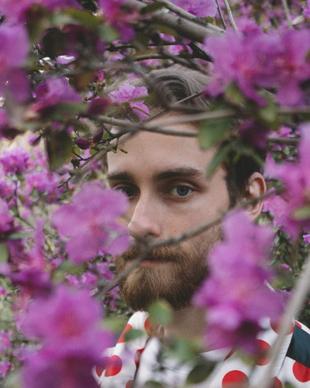 um homem com uma barba em pé na frente de flores roxas