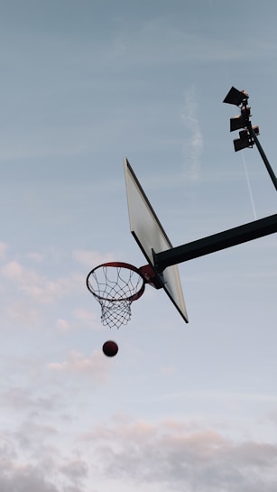 A basketball is captured in mid-air as it approaches a hoop. The scene is set against a backdrop of a clear sky, with a few clouds scattered across. The angle of the shot emphasizes the height and motion, conveying a dynamic yet peaceful atmosphere.