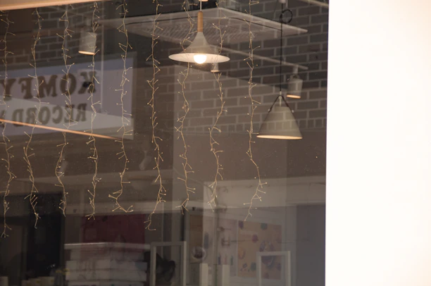 A reflection on a glass window showing interior pendant lights, fairy lights hanging down, and brick wall patterns in the background. The word 'comedy' appears to be partially visible as a reflection.