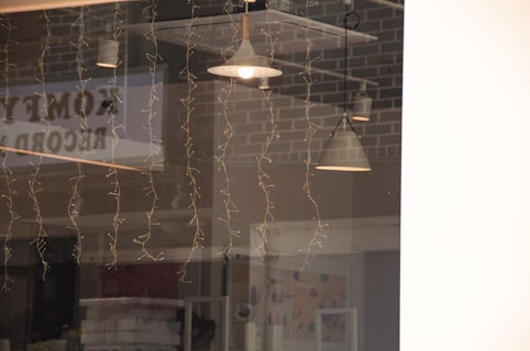 A reflection on a glass window showing interior pendant lights, fairy lights hanging down, and brick wall patterns in the background. The word 'comedy' appears to be partially visible as a reflection.
