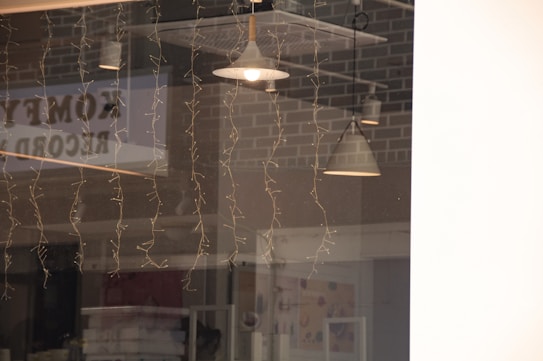 A reflection on a glass window showing interior pendant lights, fairy lights hanging down, and brick wall patterns in the background. The word 'comedy' appears to be partially visible as a reflection.