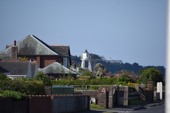 Residential area with a mix of red-brick and grey-roofed houses surrounded by green vegetation. A lighthouse is visible in the distance against a clear blue sky. The foreground includes lush greenery and a street sign partially obscured by foliage.