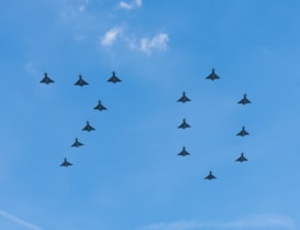 A formation of fighter jets is flying in a triangular pattern against a clear blue sky. The aircraft are evenly spaced and silhouetted against the bright background.