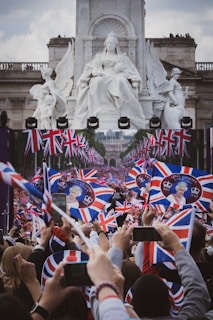 A large gathering of people enthusiastically waving Union Jack flags with images of Queen Elizabeth II. A grand statue of a seated figure with angels is in the background, creating a prominent and majestic scene. The crowd is capturing the event with smartphones.