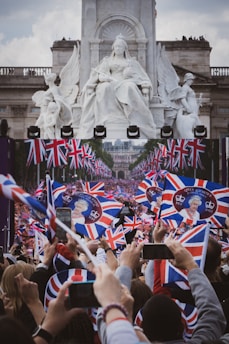 A large gathering of people enthusiastically waving Union Jack flags with images of Queen Elizabeth II. A grand statue of a seated figure with angels is in the background, creating a prominent and majestic scene. The crowd is capturing the event with smartphones.