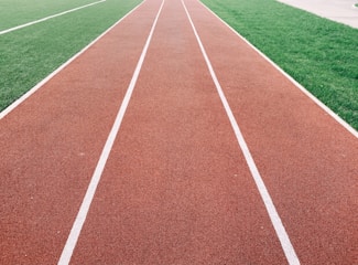 A red running track with white lane markings is adjacent to a lush green field. The linear perspective of the track draws the eye into the distance.