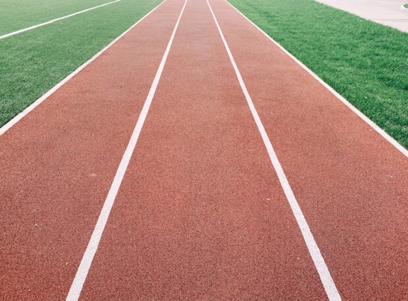 A red running track with white lane markings is adjacent to a lush green field. The linear perspective of the track draws the eye into the distance.