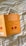 A book with an orange cover featuring the title in German by John Gray, next to an image partially tucked inside it. The background is a white fabric with yellow and gray stripes.