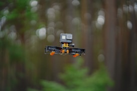 A small drone hovers in mid-air, equipped with a camera mounted on top. The background features a blurred forest, indicating the drone is flying outdoors. The drone has visible propellers and a compact design.