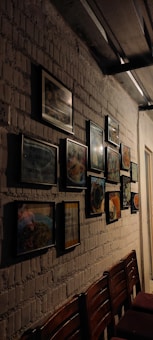 A dimly lit interior featuring a white brick wall adorned with several framed pictures, possibly of food or quotes. Below the frames, there are wooden chairs aligned along the wall under soft lighting.
