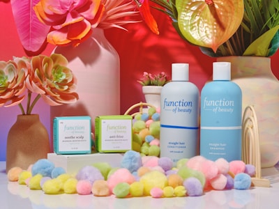 A colorful arrangement featuring skincare products, including bottles labeled 'function of beauty' for shampoo and conditioner. Small boxes for haircare booster shots are also present. The scene is accentuated with vibrant flowers in vases and pastel-colored pom-poms scattered around.