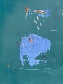 A large patch of blue paint is peeling off a surface with a green undercoat, revealing rust spots and minor scratches. The texture of the surface is rough and weathered, suggesting exposure to natural elements over time.