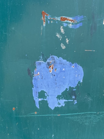 A large patch of blue paint is peeling off a surface with a green undercoat, revealing rust spots and minor scratches. The texture of the surface is rough and weathered, suggesting exposure to natural elements over time.