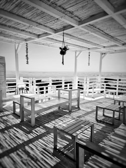A wooden terrace with a pergola roof casts patterned shadows on the floor. Hanging decor and minimalist wooden benches and tables create a relaxed, open space overlooking a distant view.