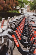 A row of rental bicycles neatly lined up along a sidewalk in an urban area. The bicycles are primarily gray with bright red branding on the frame. In the background, there are trees and residential buildings, creating a serene street scene.