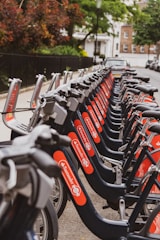 A row of rental bicycles neatly lined up along a sidewalk in an urban area. The bicycles are primarily gray with bright red branding on the frame. In the background, there are trees and residential buildings, creating a serene street scene.