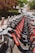 A row of rental bicycles neatly lined up along a sidewalk in an urban area. The bicycles are primarily gray with bright red branding on the frame. In the background, there are trees and residential buildings, creating a serene street scene.