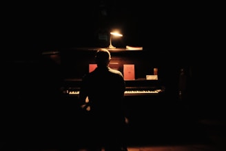 A person is seated at an upright piano in a dimly lit room. A small lamp casts light over the piano, illuminating a few objects and sheet music on the piano's surface. The atmosphere is serene and focused.