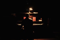 A person is seated at an upright piano in a dimly lit room. A small lamp casts light over the piano, illuminating a few objects and sheet music on the piano's surface. The atmosphere is serene and focused.