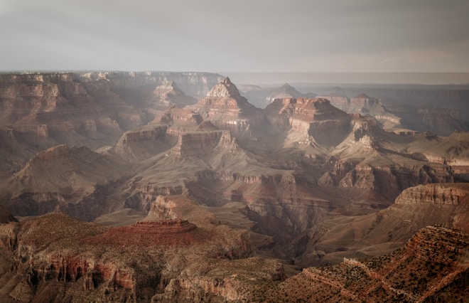 An expansive view of the Grand Canyon, showcasing rugged cliffs and deep valleys with layers of rock in varying shades of brown, red, and beige. The landscape is vast and dramatic, with a hazy sky overhead adding a sense of depth and scale to the scene.