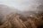 A vast, rugged landscape of the Grand Canyon with layers of rock formations and cliffs extending into the distance. The scene is enveloped in a light haze, with muted earth tones dominating the view.