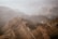 A vast, rugged landscape of the Grand Canyon with layers of rock formations and cliffs extending into the distance. The scene is enveloped in a light haze, with muted earth tones dominating the view.
