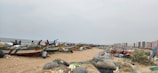 A stretch of sandy beach with several fishing boats lined up, filled with bundled nets. People can be seen preparing or attending to the boats. In the background, there are rows of buildings, likely residential, providing a contrast between the natural and urban environment. Various fishing nets and supplies are scattered across the sand.