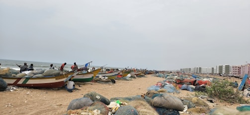 A stretch of sandy beach with several fishing boats lined up, filled with bundled nets. People can be seen preparing or attending to the boats. In the background, there are rows of buildings, likely residential, providing a contrast between the natural and urban environment. Various fishing nets and supplies are scattered across the sand.