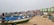 A sandy beach is lined with colorful fishing boats, nets, and various fishing equipment. Several people, including fishermen, are seen attending to their tasks. In the background, there are modern apartment buildings with vibrant green and pink colors, stretching along the coastline under an overcast sky.