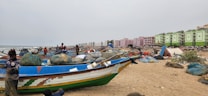 A sandy beach is lined with colorful fishing boats, nets, and various fishing equipment. Several people, including fishermen, are seen attending to their tasks. In the background, there are modern apartment buildings with vibrant green and pink colors, stretching along the coastline under an overcast sky.