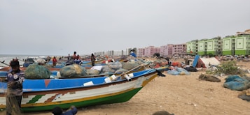 A sandy beach is lined with colorful fishing boats, nets, and various fishing equipment. Several people, including fishermen, are seen attending to their tasks. In the background, there are modern apartment buildings with vibrant green and pink colors, stretching along the coastline under an overcast sky.