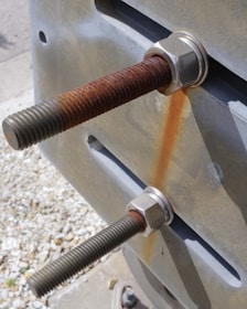 Two large metal bolts with nuts are fastened onto a metal surface, exhibiting visible rust on one of the bolts. The background features pebbled ground with soft focus.