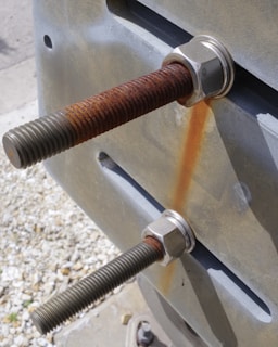 Two large metal bolts with nuts are fastened onto a metal surface, exhibiting visible rust on one of the bolts. The background features pebbled ground with soft focus.