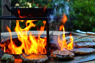 A close-up view of a grill with sizzling steaks cooking over an open flame. The vibrant fire emits bright orange and yellow hues, casting a warm glow. Wisps of smoke rise from the sizzling meat, adding to the rustic outdoor cooking atmosphere. In the background, blurred greenery provides a natural setting.