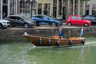 A small wooden boat navigates a calm canal, with a person steering. The canal is lined with parked cars and old buildings, reflecting an urban setting. A flag is attached to the boat, indicating a cultural or national symbol.