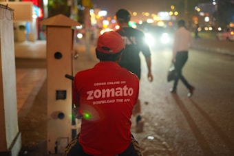 A person wearing a red Zomato delivery shirt and cap is sitting on a motorcycle or scooter on a dimly lit street. The background shows blurred lights, possibly from cars and streetlights, creating a bokeh effect. Other individuals are seen walking in the dimly lit street.