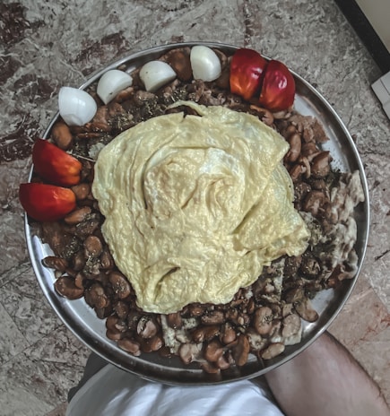 A large metal platter filled with a mix of cooked brown beans and topped with a large, folded yellow omelette. Surrounding the beans are several pieces of sliced red and white onions, as well as halved red bell peppers. The platter is placed on a marbled surface.