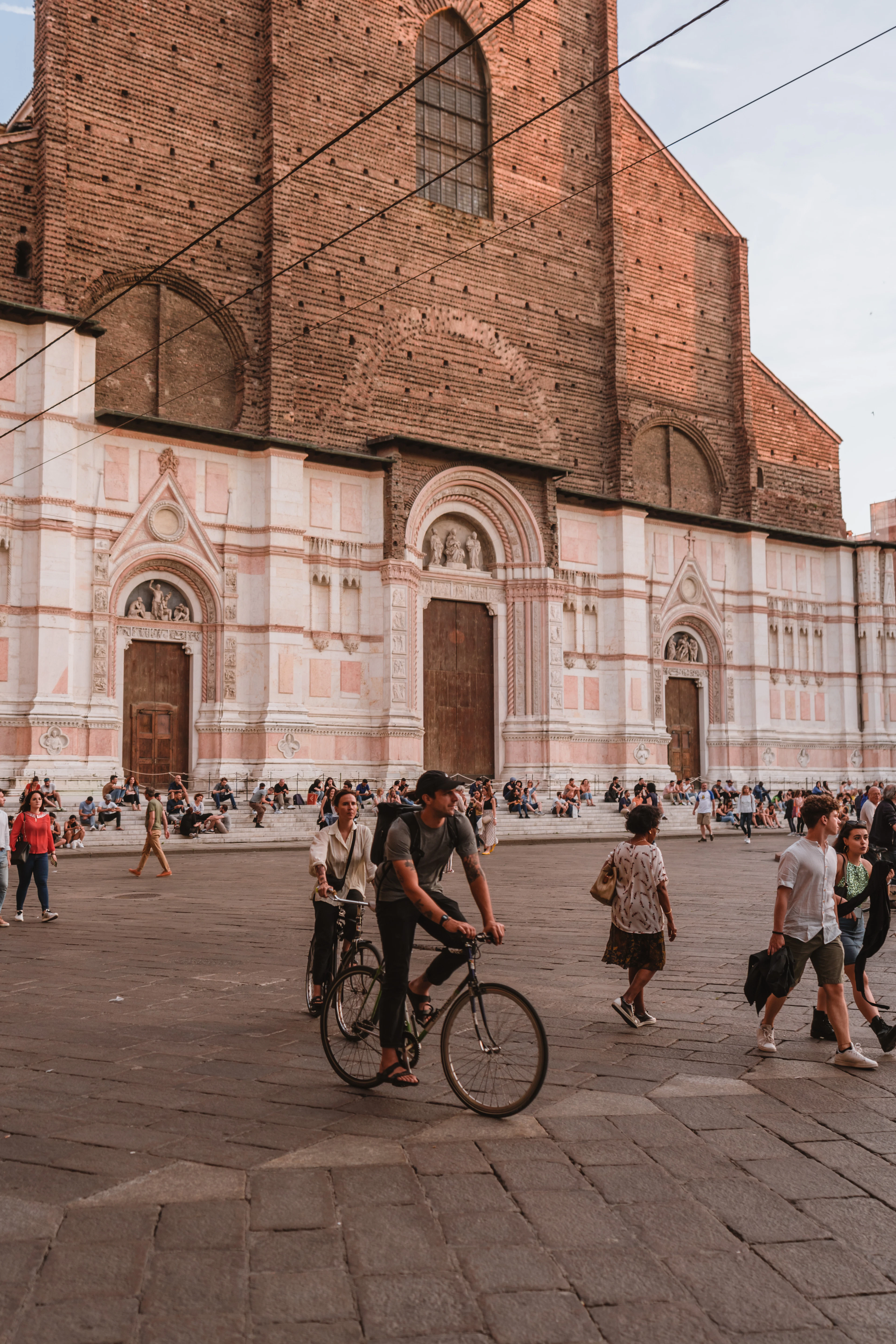 A cyclist passes the brick and pink-stone facade of Bologna's Basilica of San Petronio at golden hour.
