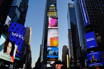 A cityscape with tall skyscrapers adorned with bright, colorful digital billboards. Various ads are displayed for well-known brands, including Yahoo, Dunkin' Donuts, and FOX. The scene is captured during the early evening, with a clear sky above.