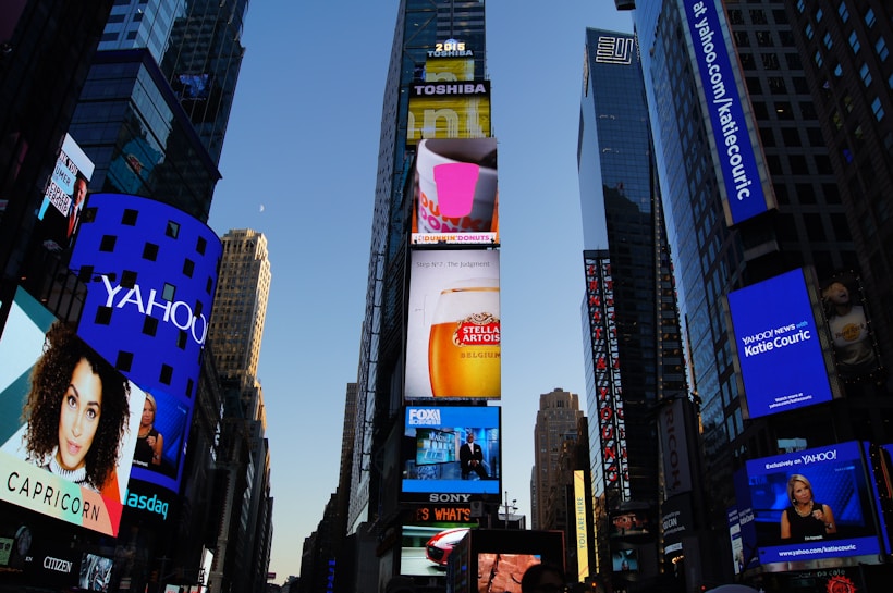 A cityscape with tall skyscrapers adorned with bright, colorful digital billboards. Various ads are displayed for well-known brands, including Yahoo, Dunkin' Donuts, and FOX. The scene is captured during the early evening, with a clear sky above.