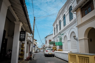 A narrow street flanked by white colonial-style buildings with arched windows and balconies. There are overhead power lines connecting across the street. A parked white car is visible along with some motorbikes near a shop displaying a sign for Ibrahim Jewellers.