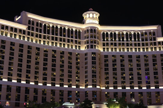 A large, well-lit hotel building with numerous windows and symmetrical architectural design features prominently against the night sky. The structure has a central tower with arches and a decorative circular top, while the lower floors include evenly spaced balconies and lighting accents that highlight its grandeur.